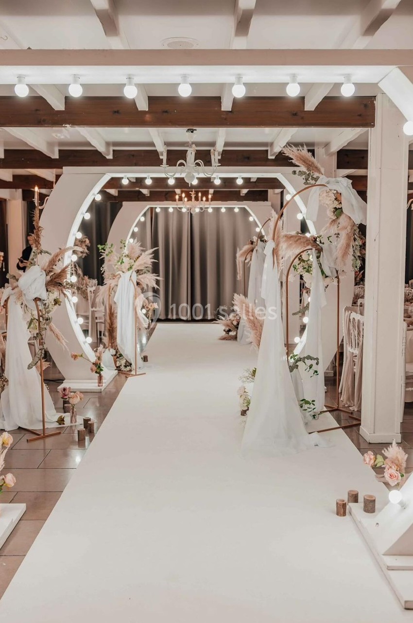 Allée décorée pour un mariage avec arches lumineuses, voilages blancs et arrangements floraux dans une salle élégante.