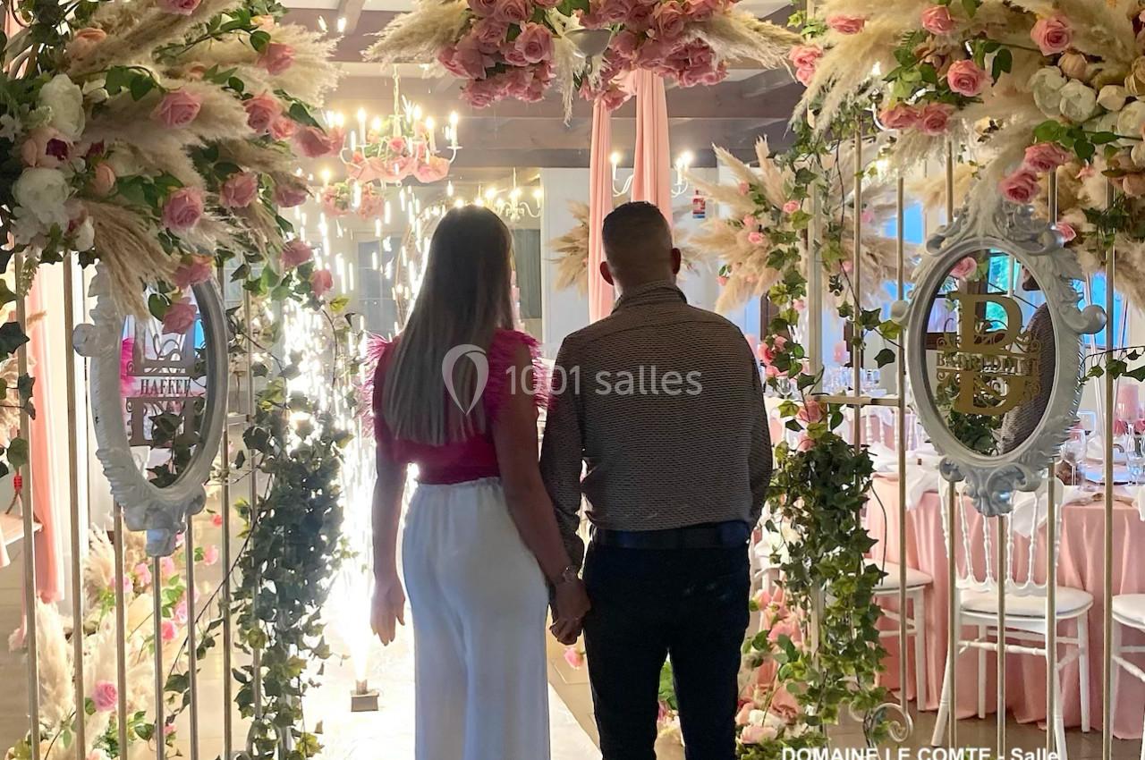 Un couple de dos traverse une arche fleurie dans une salle décorée de roses et de lumières.