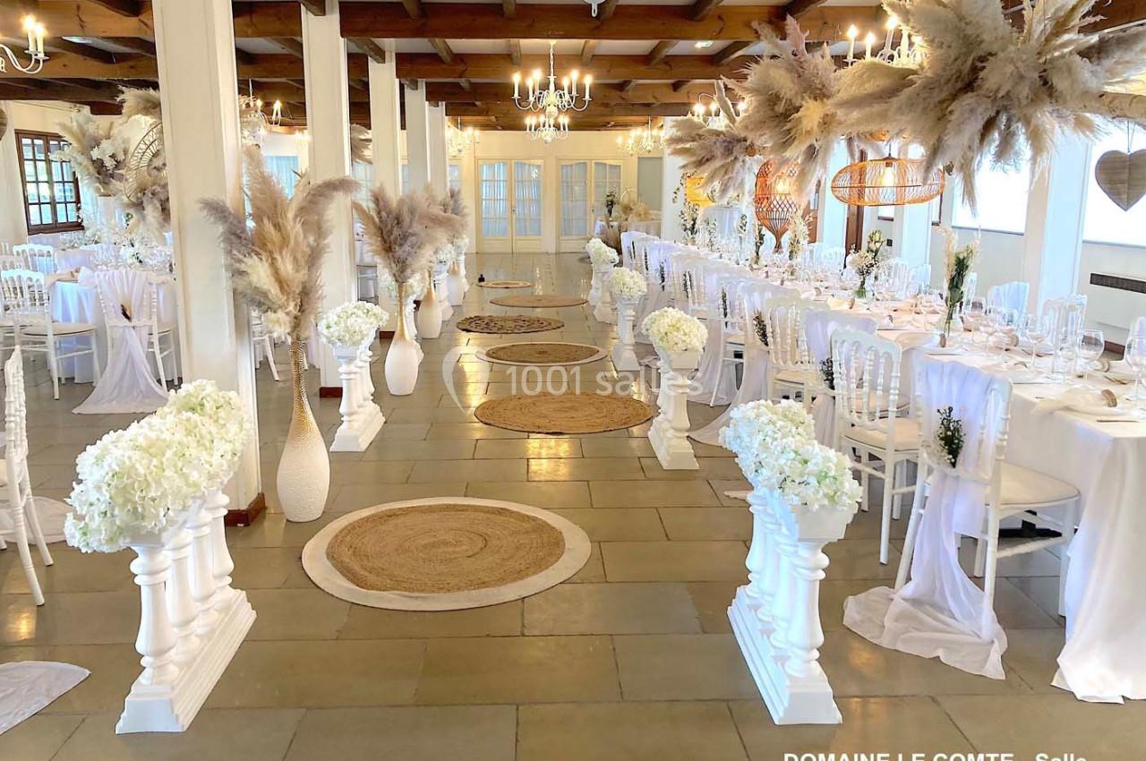 Salle de réception lumineuse décorée avec des fleurs blanches, des pampas et des chaises blanches autour de tables dressées.