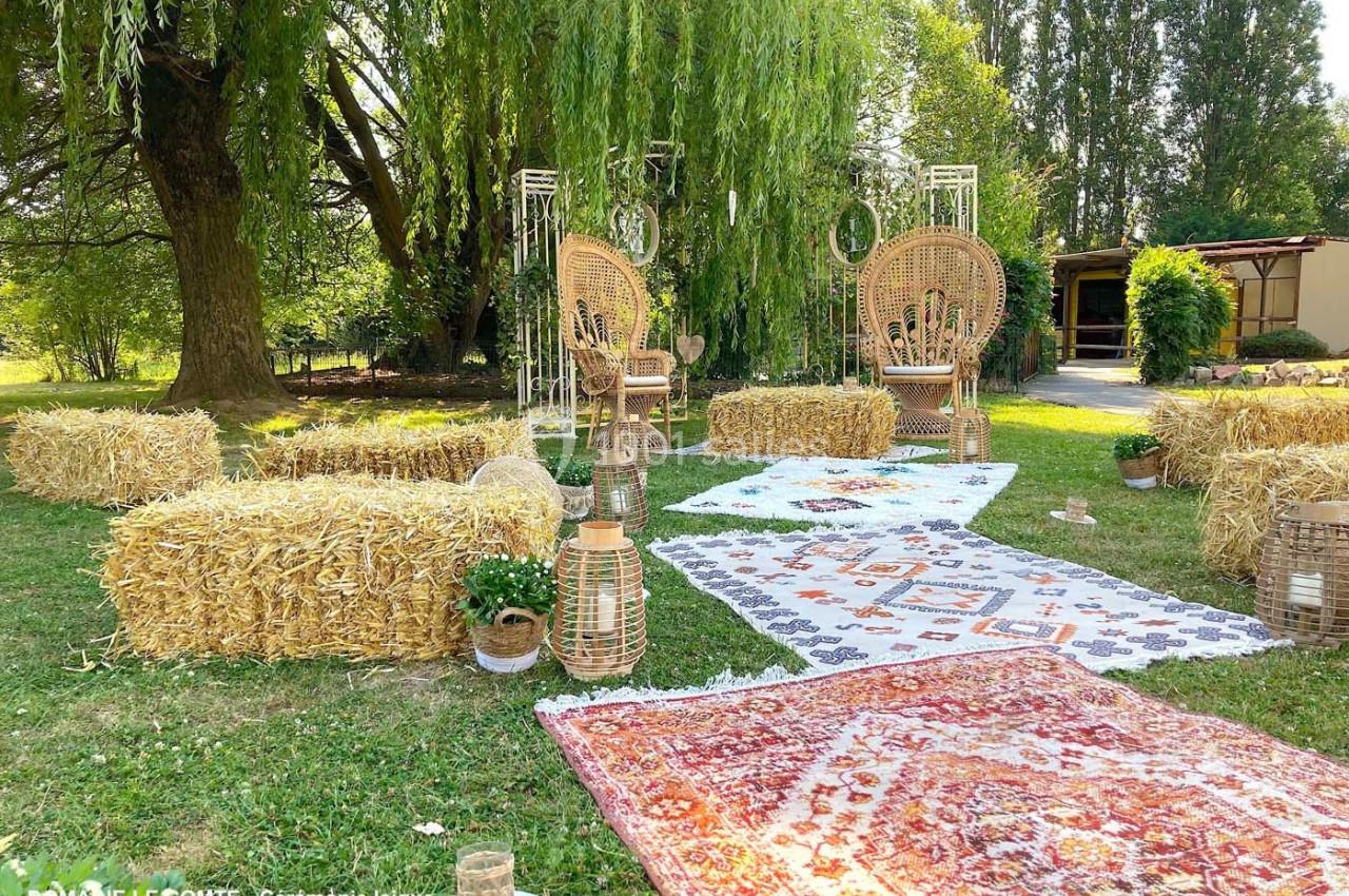 Décor champêtre avec tapis colorés, bottes de paille, fauteuils en osier et lanternes sous un grand arbre.