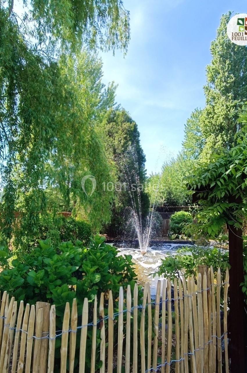 Un jardin verdoyant avec une fontaine centrale entourée de végétation et une clôture en bois au premier plan.