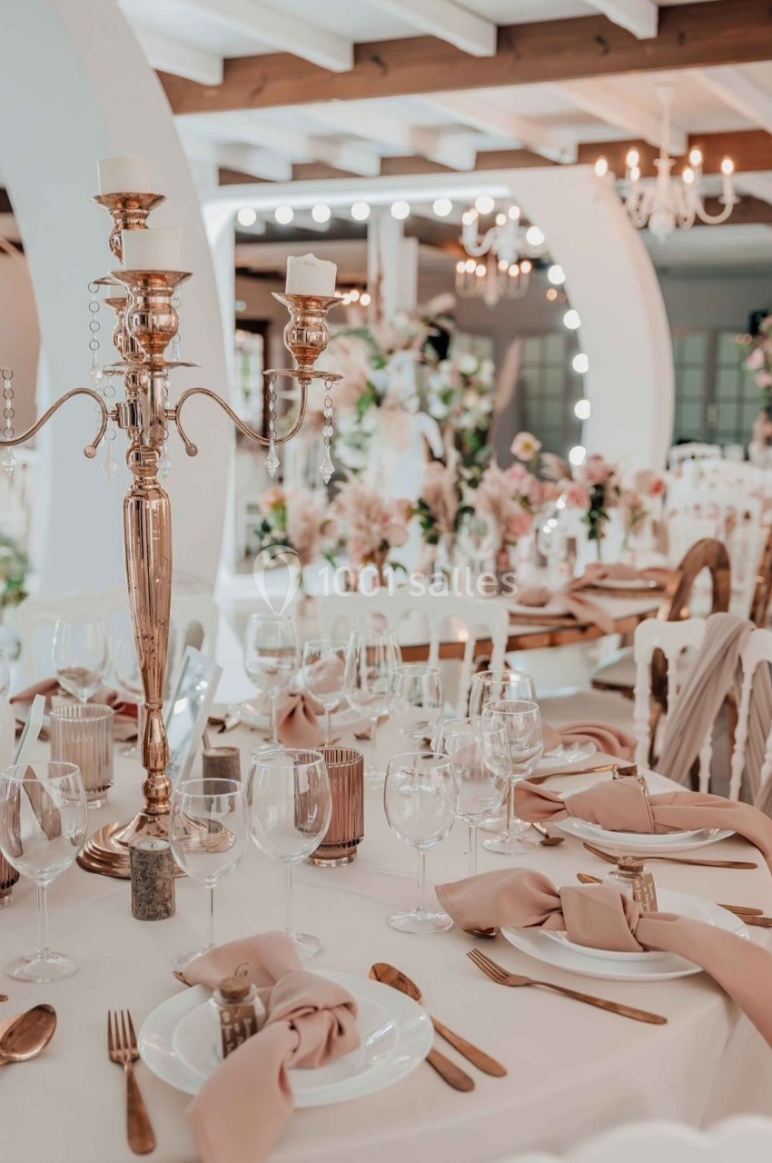 Salle de réception élégante avec tables décorées de nappes blanches, chandeliers dorés et serviettes roses.
