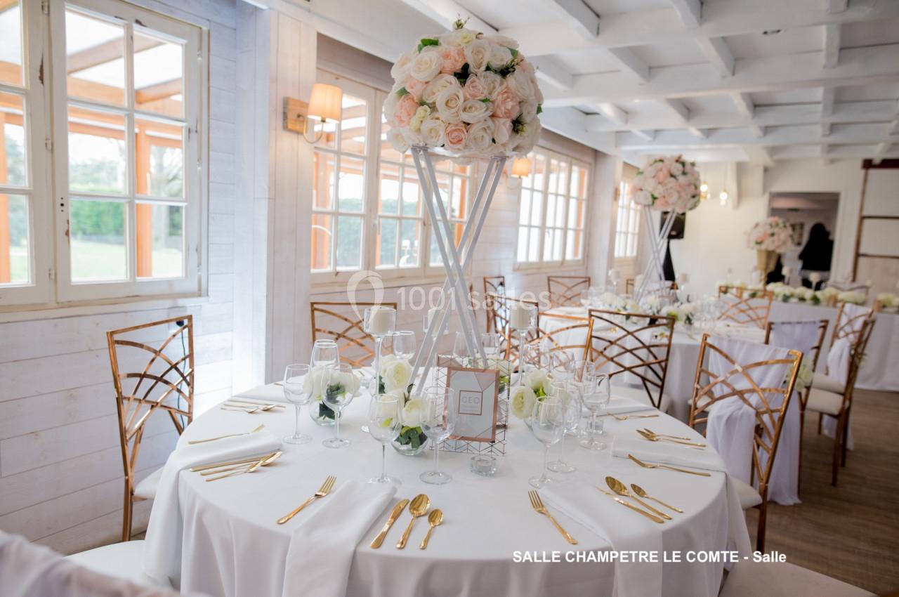 Salle de réception lumineuse avec tables rondes décorées de nappes blanches, fleurs et couverts dorés.