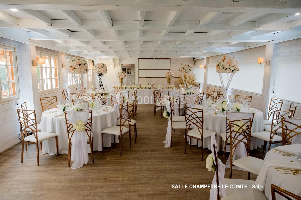 Salle de réception lumineuse avec tables rondes décorées de nappes blanches, chaises en bois et arrangements floraux.