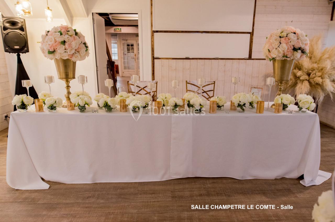 Table décorée avec des bouquets de fleurs blanches et roses, chandeliers dorés et nappes blanches dans une salle rustique.