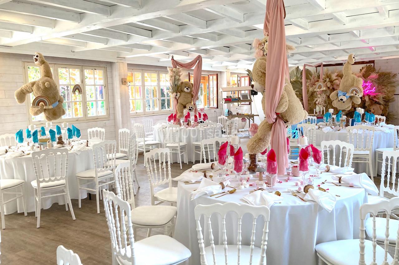 Salle de réception décorée avec des tables rondes, nappes blanches, serviettes colorées et peluches suspendues.