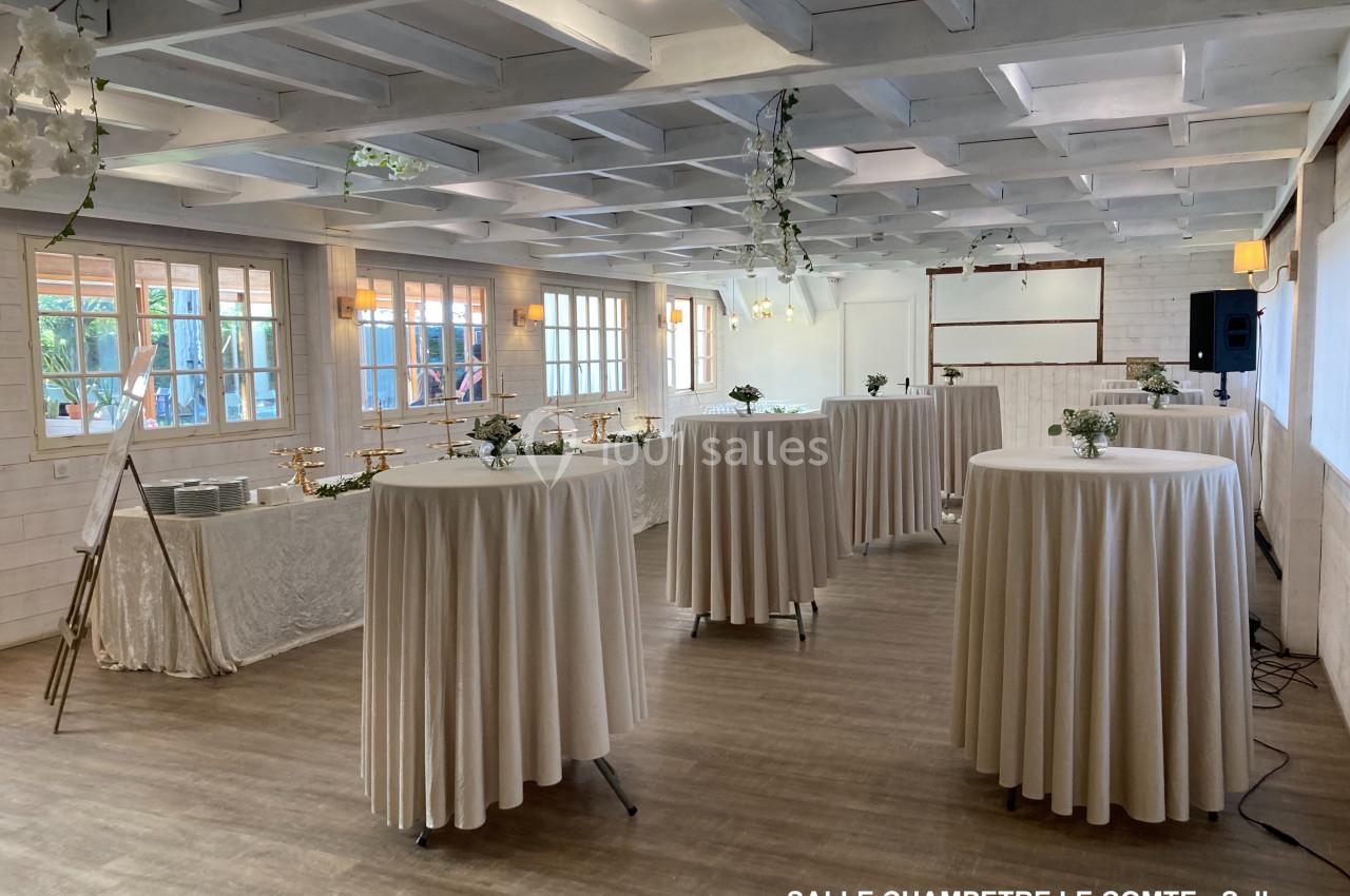 Salle lumineuse avec tables hautes drapées de blanc, décorée de fleurs, parquet au sol et poutres apparentes au plafond.