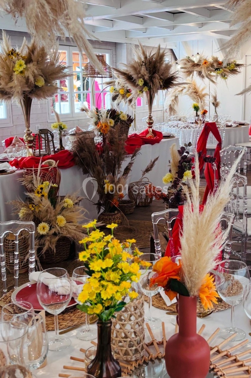 Salle décorée pour un événement avec des tables ornées de fleurs séchées, vases colorés et nappes rouges.