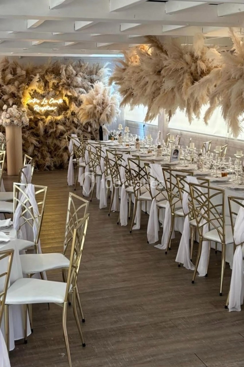 Salle de réception décorée avec des pampas, tables dressées avec nappes blanches et chaises ornées de rubans.