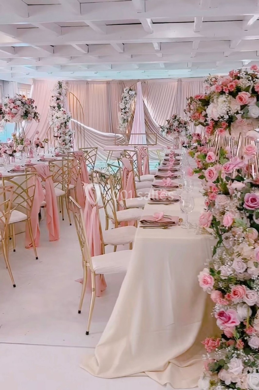 Salle de réception décorée avec des tables élégantes, nappes blanches et arrangements floraux roses et blancs.