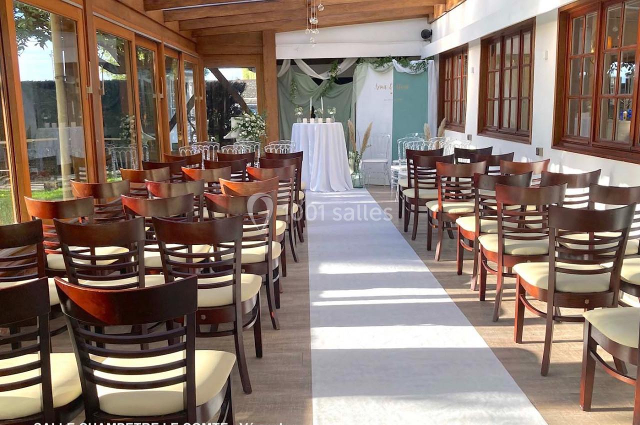 Salle de réception lumineuse avec chaises en bois disposées en rangées, allée centrale et table décorée au fond.