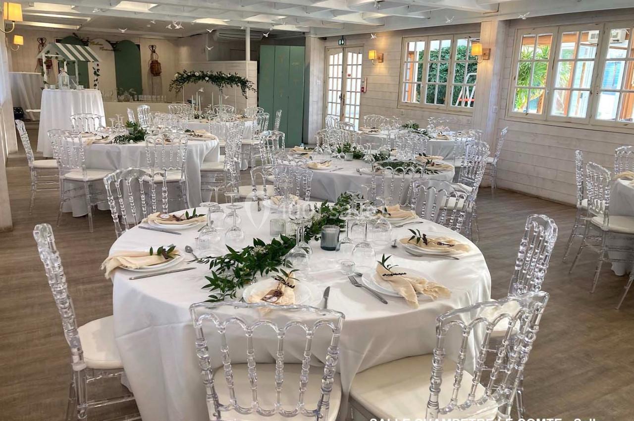 Salle décorée pour un événement, avec tables rondes dressées, nappes blanches, chaises transparentes et ornements végétaux.