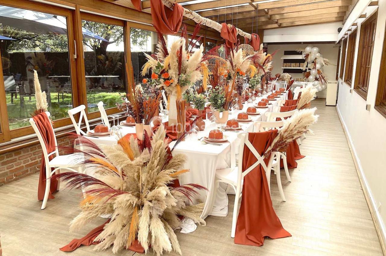 Salle décorée pour un événement avec tables alignées, nappes blanches, chemins de table orange et ornements de fleurs…