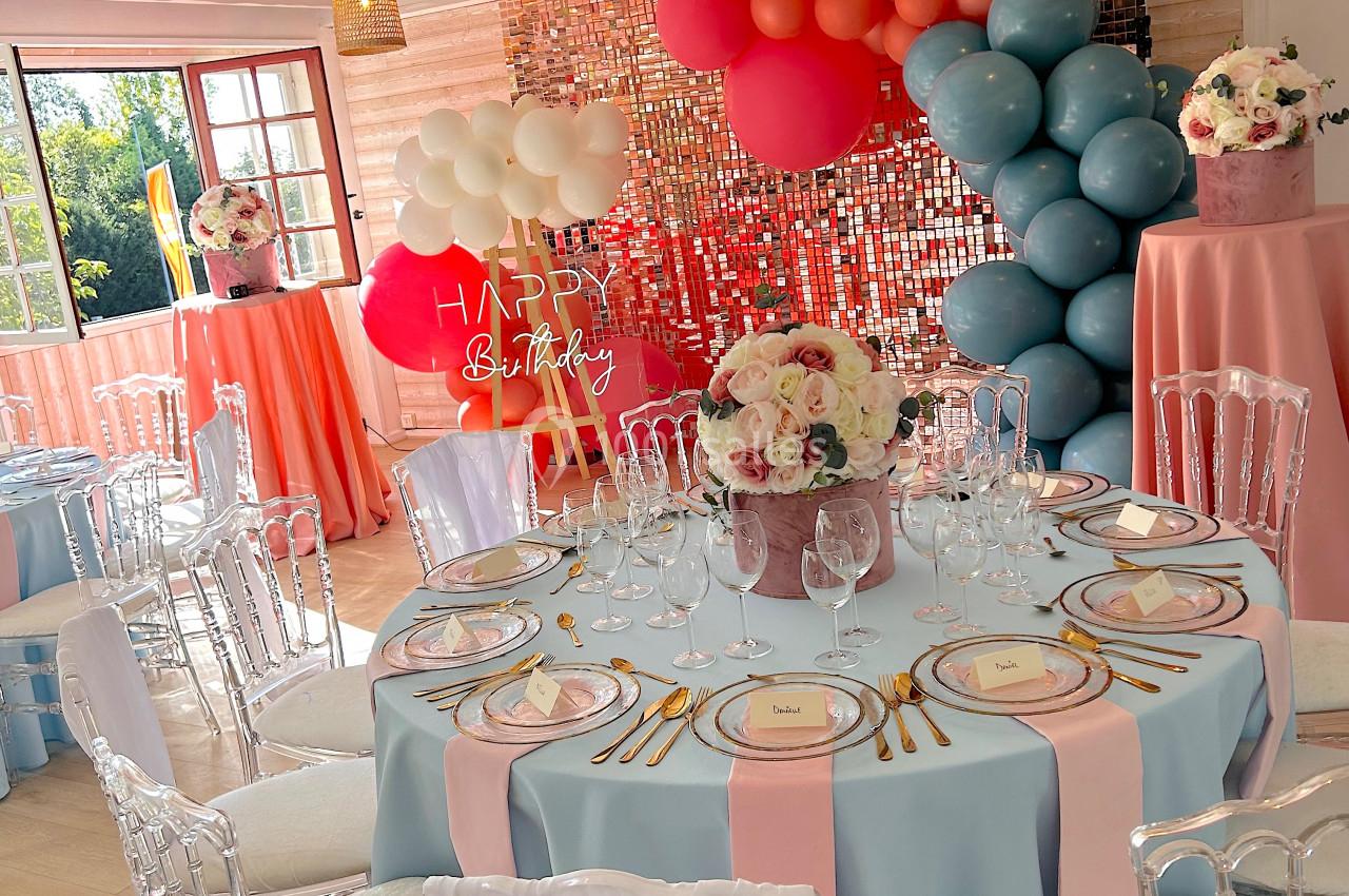 Salle décorée pour une fête avec des tables dressées, des ballons colorés et un mur scintillant en arrière-plan.