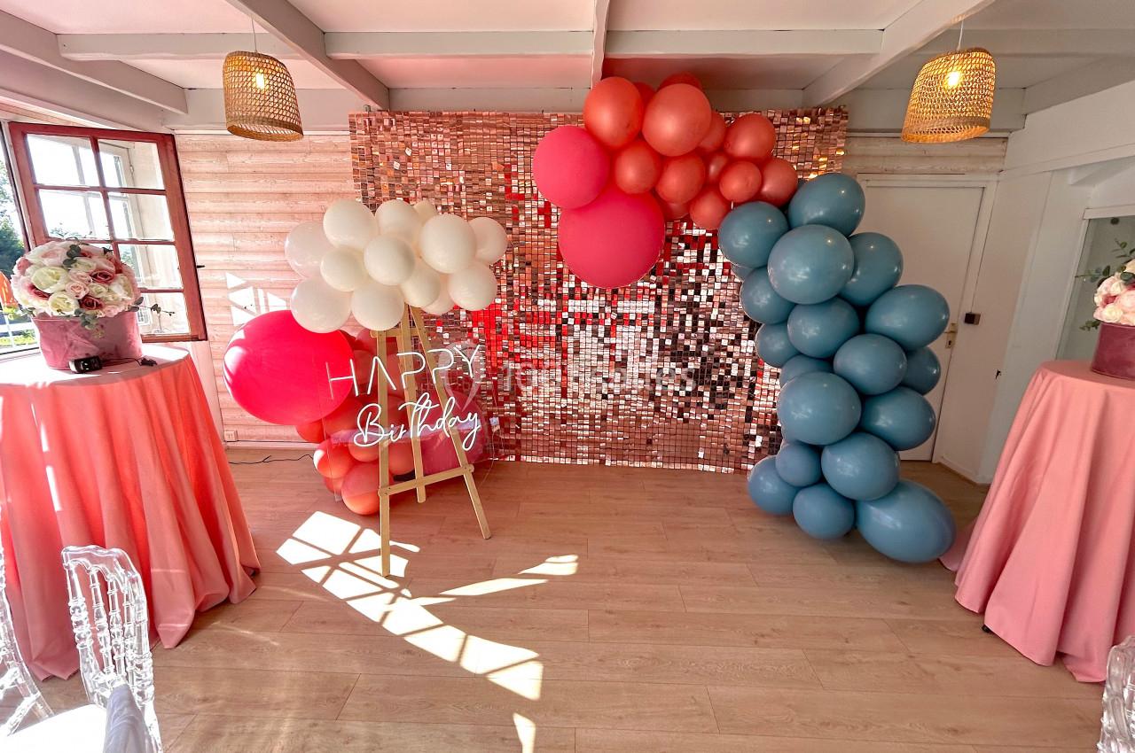 Décoration d'anniversaire avec ballons colorés, mur scintillant, panneau ’Happy Birthday’ et tables drapées de tissu rose.