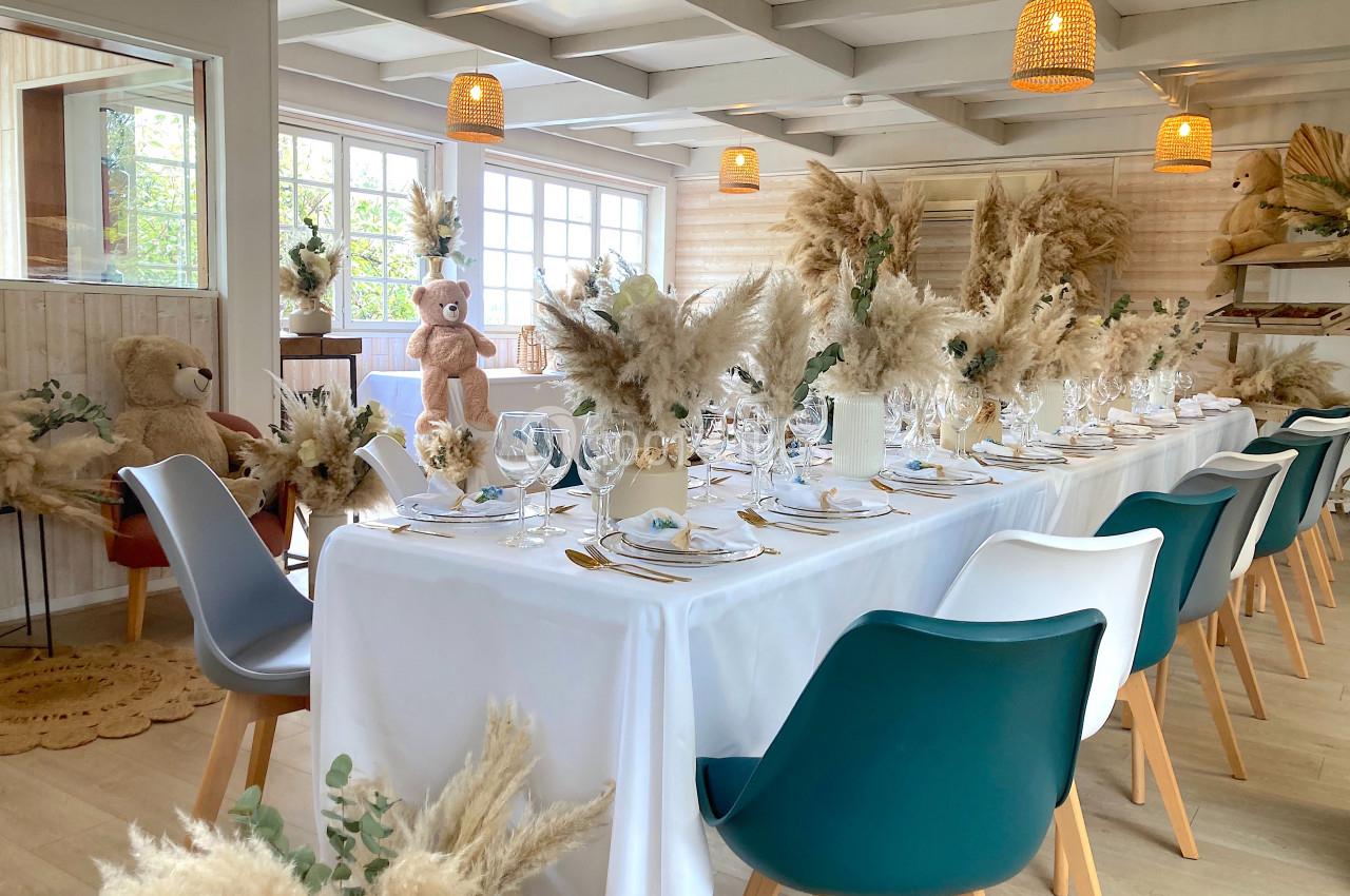 Salle décorée pour un événement avec une table dressée, des chaises colorées et des arrangements de fleurs séchées.
