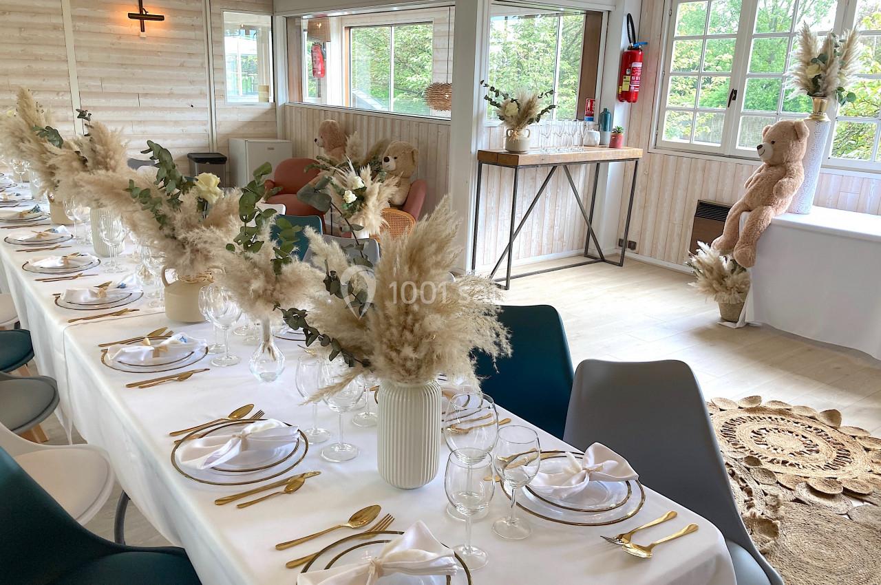 Salle lumineuse décorée pour un événement, avec une longue table dressée, des fleurs séchées et des ours en peluche.