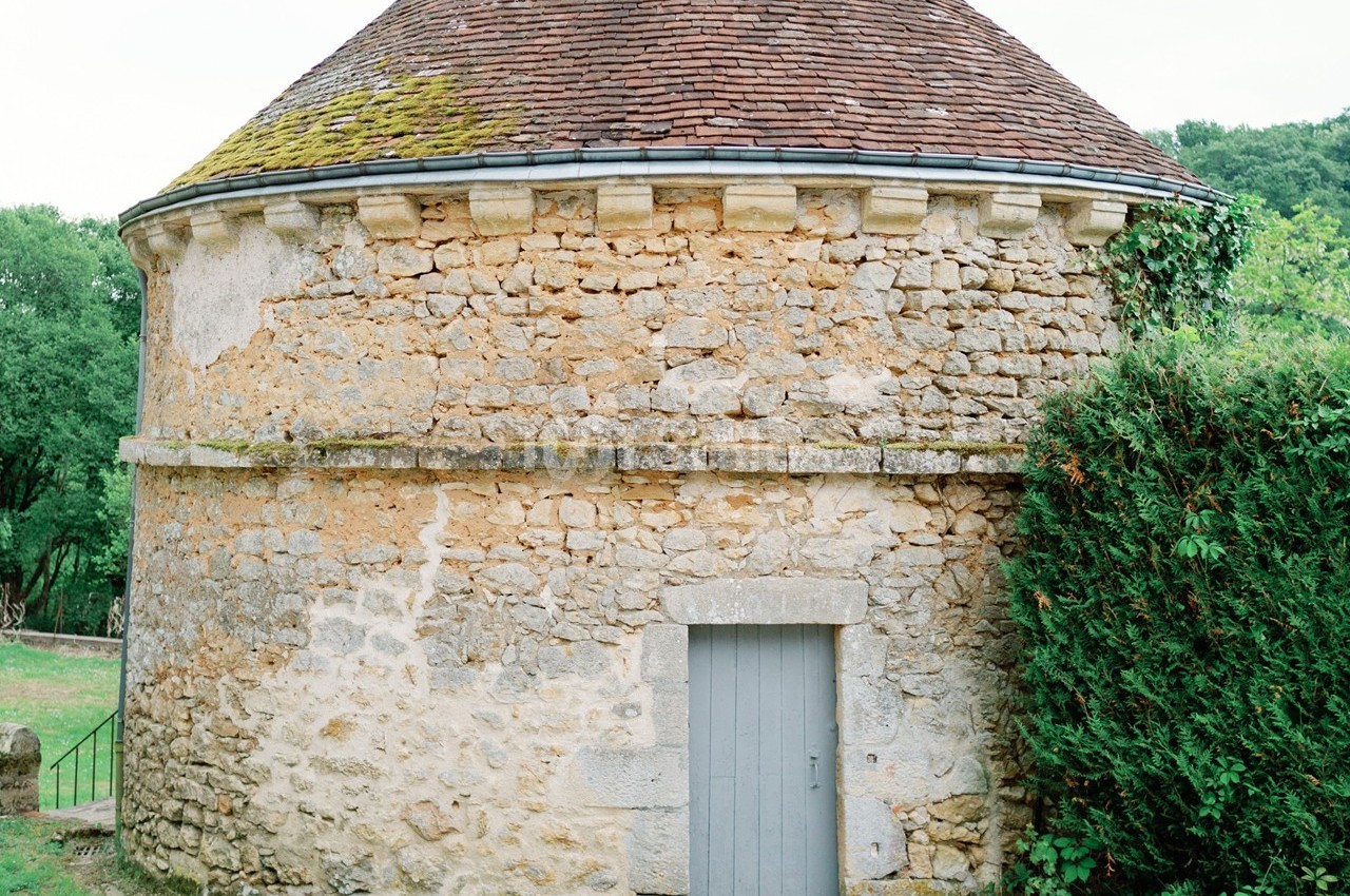 Bâtiment en pierre circulaire avec un toit en tuiles, une porte grise et de la végétation à droite.