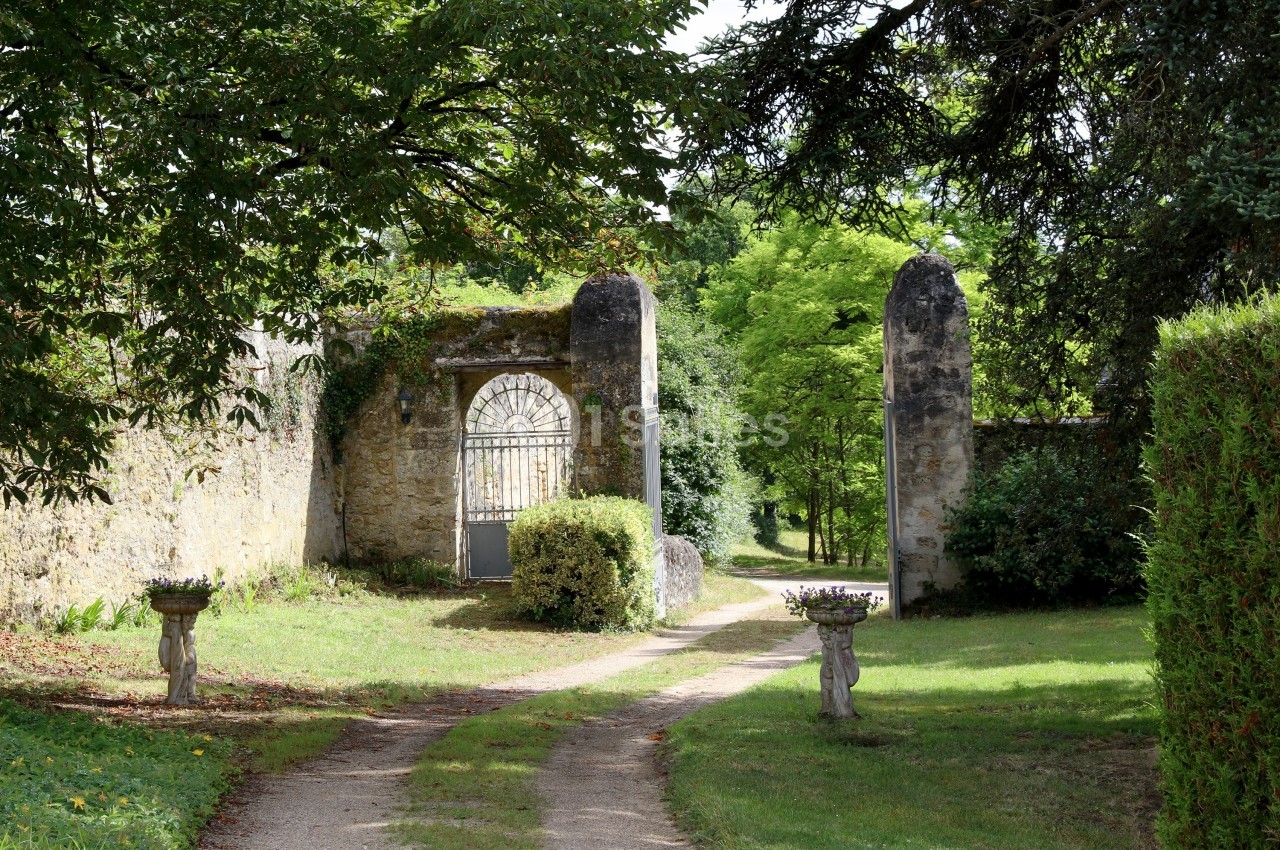 Allée bordée de verdure menant à une porte en fer forgé encadrée par des piliers en pierre dans un jardin.