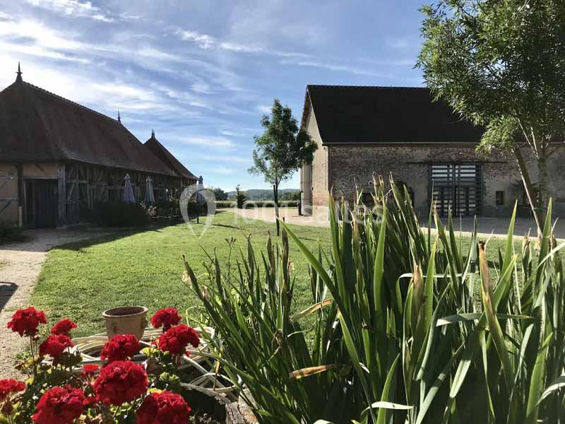 Cour d'une ferme rénovée avec pelouse, fleurs rouges au premier plan, bâtiments en pierre et ciel dégagé.