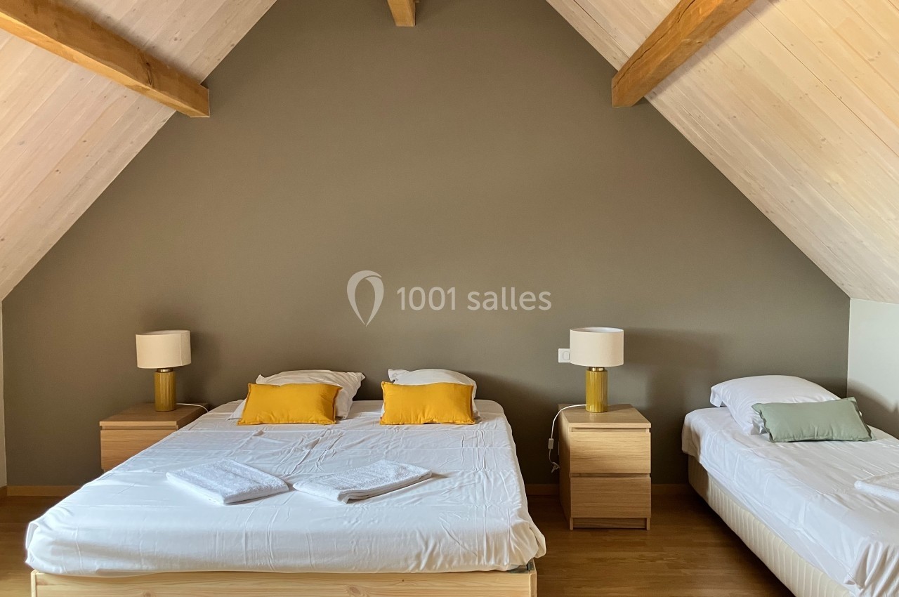 Chambre mansardée avec un lit double, un lit simple, des tables de chevet en bois et des lampes, sur un sol en parquet.
