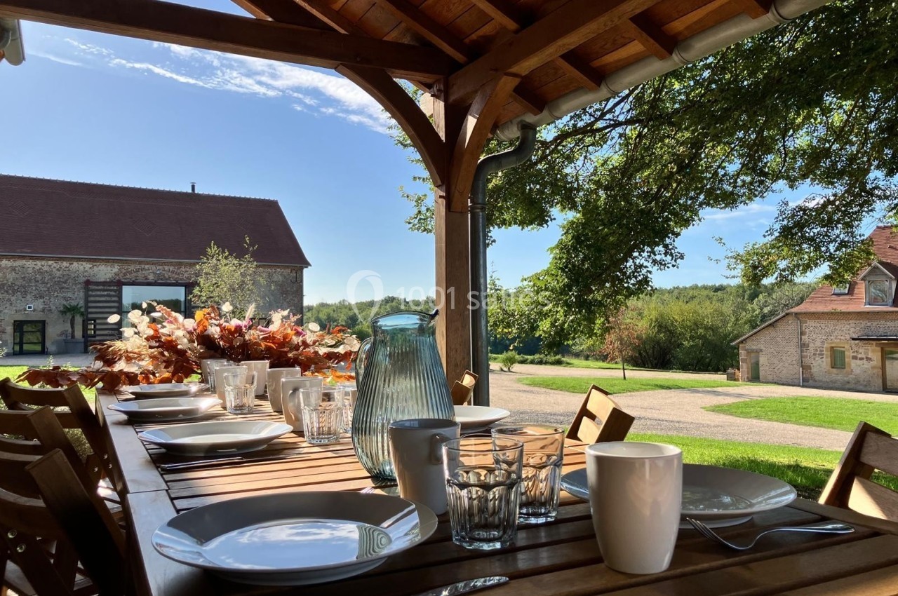 Table en bois dressée avec vaisselle et carafe, sous une pergola, donnant sur un jardin et des bâtiments ruraux.