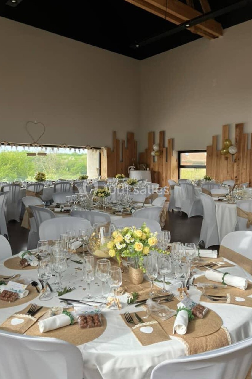 Salle de réception décorée pour un mariage, avec tables rondes dressées, nappes blanches et centres de table fleuris.