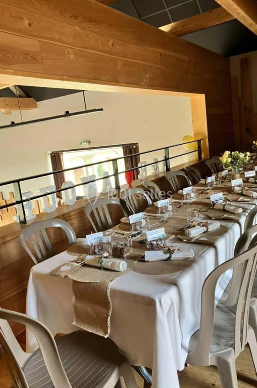 Table dressée avec nappes blanches, couverts, décorations rustiques et chaises dans une salle en bois lumineuse.