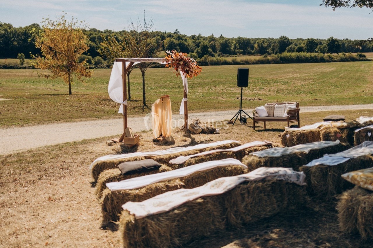 Cérémonie en plein air avec des bottes de foin en guise de sièges et une arche décorée de fleurs dans un champ.