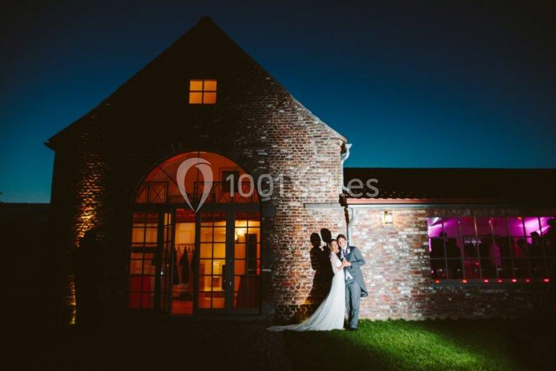 Un couple en tenue de mariage pose devant un bâtiment en briques éclairé à la tombée de la nuit.