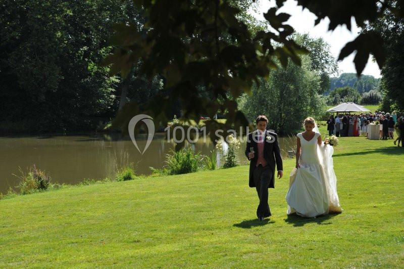 Un couple en tenue de mariage marche sur une pelouse près d'un étang, avec des invités rassemblés au loin.