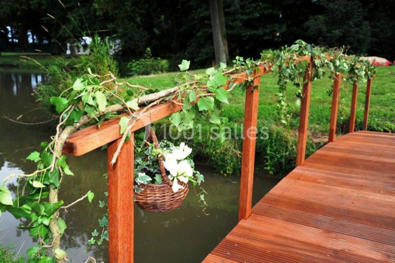 Petit pont en bois décoré de feuillages et d'un panier fleuri, traversant un cours d'eau dans un cadre verdoyant.