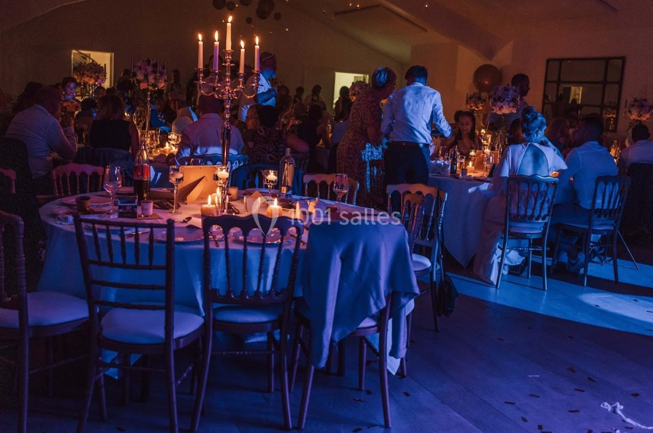 Salle de réception éclairée par des bougies, avec des tables décorées et des invités assis ou debout.