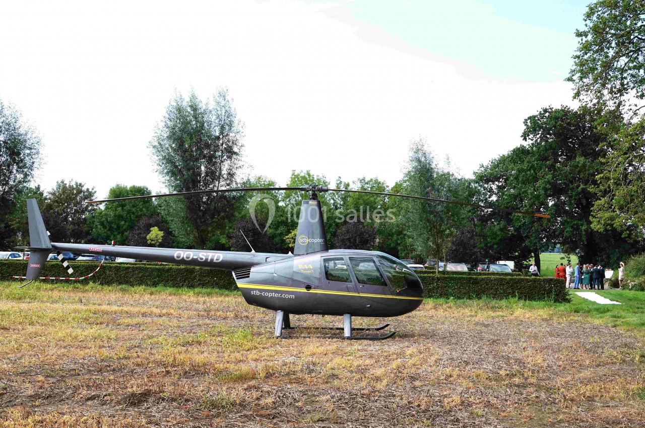 Hélicoptère gris posé sur un terrain herbeux, entouré d'arbres et de quelques personnes en arrière-plan.