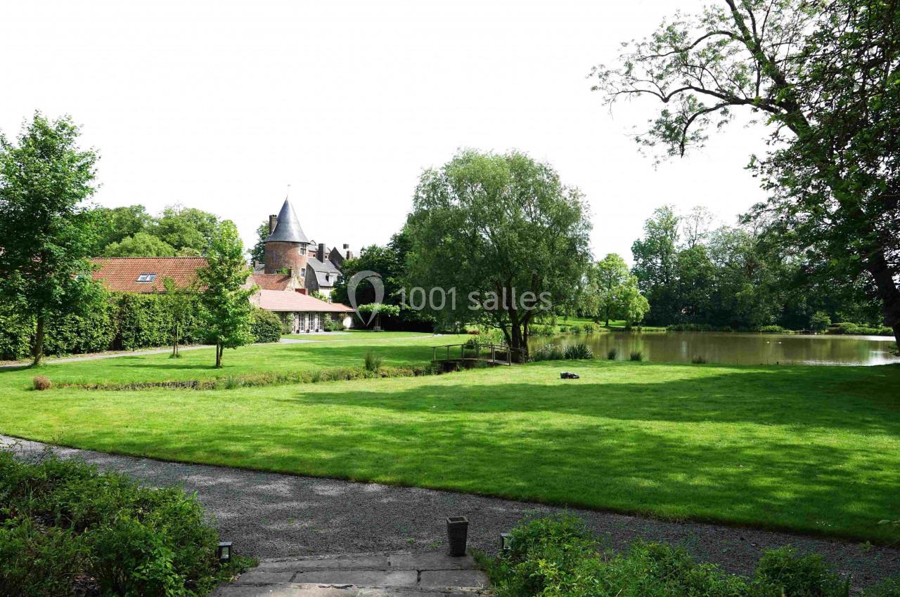 Vue d'un jardin verdoyant avec pelouse, arbres, un étang et une bâtisse en arrière-plan surmontée d'une tour.