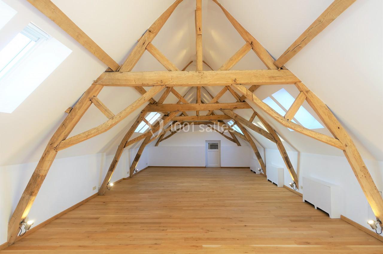 Combles aménagés avec poutres apparentes en bois, parquet clair et lumière naturelle provenant de fenêtres de toit.