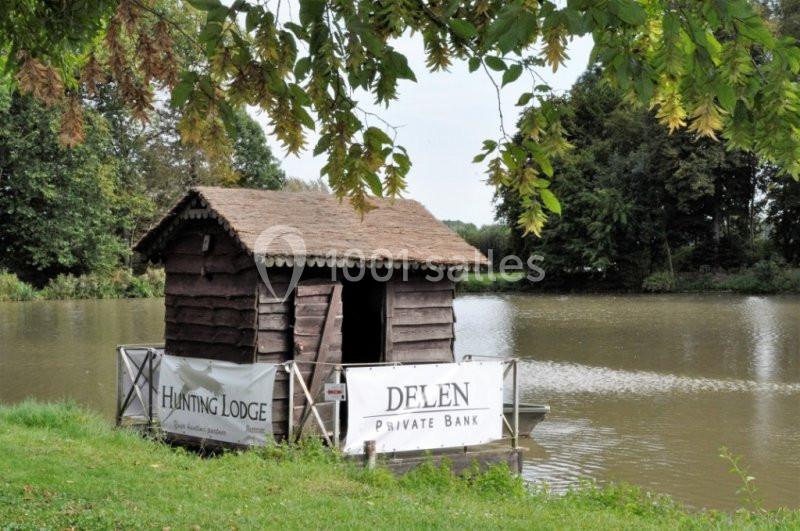 Cabane en bois sur pilotis au bord d'un étang, entourée de végétation et partiellement décorée de banderoles.