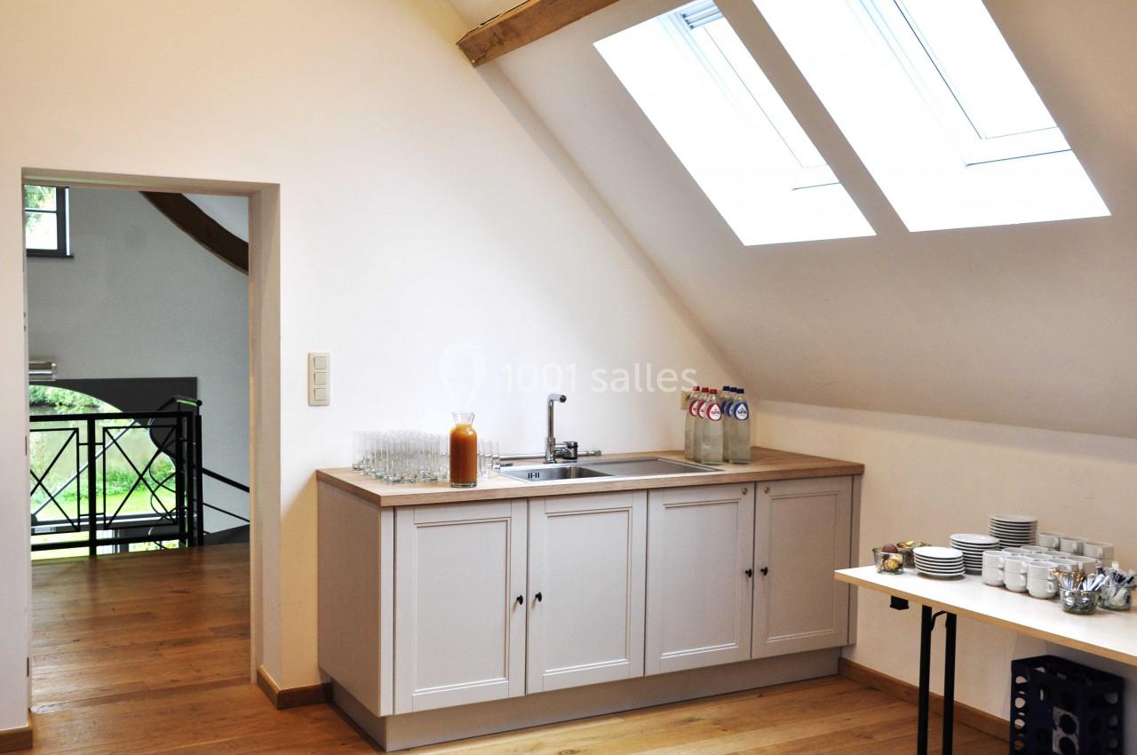 Cuisine lumineuse sous combles avec évier, bouteilles d'eau, verres et vaisselle sur un plan de travail en bois.