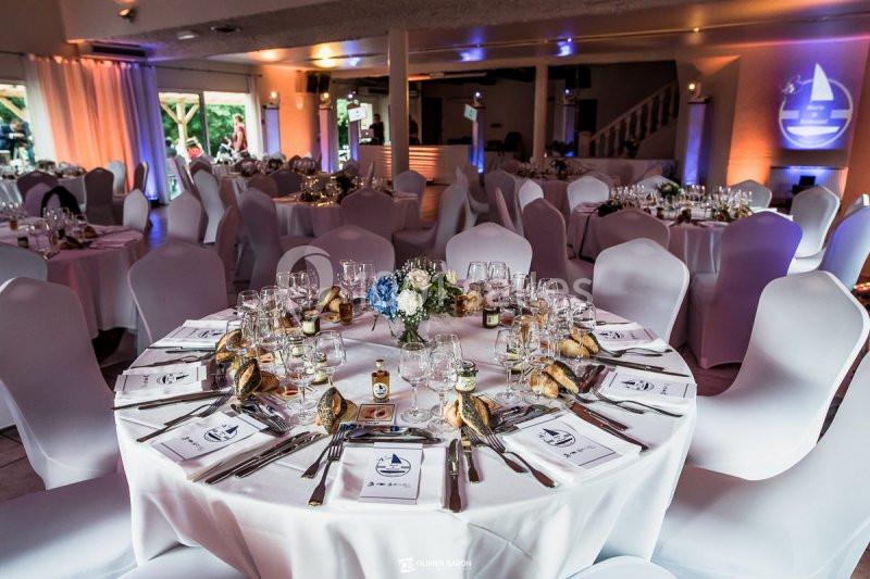 Salle de réception avec tables rondes dressées, nappes blanches, couverts élégants et décoration florale au centre.