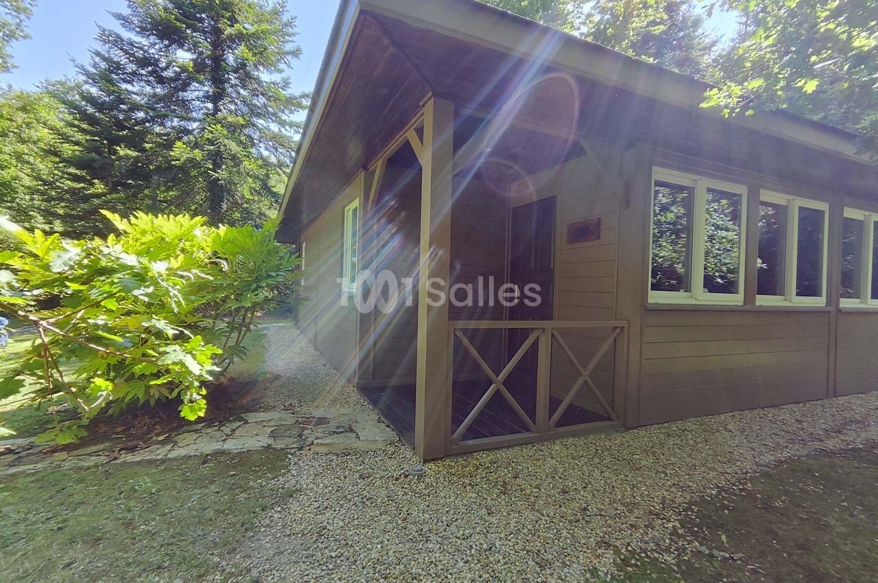 Chalet en bois entouré de végétation, avec une allée de gravier et des rayons de soleil traversant les arbres.