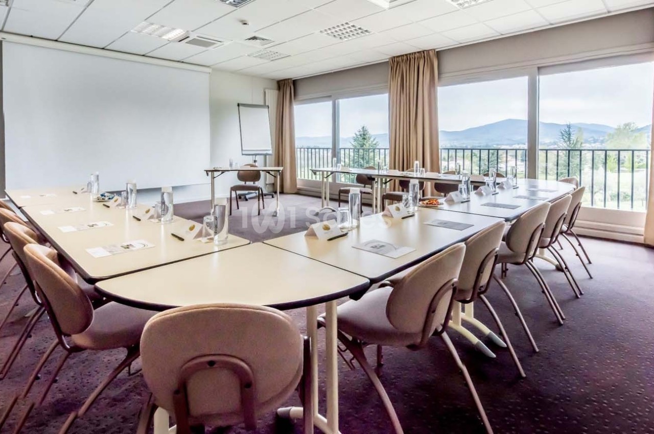 Salle de réunion lumineuse avec tables disposées en U, chaises, paperboards et vue sur un paysage extérieur.
