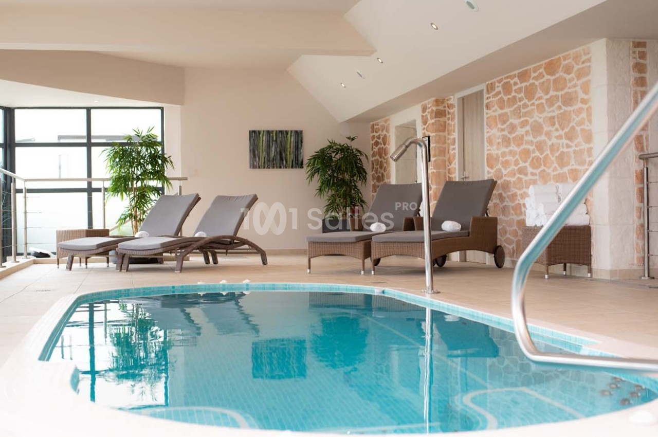 Piscine intérieure avec transats, plantes et murs en pierre, éclairée par de grandes fenêtres.