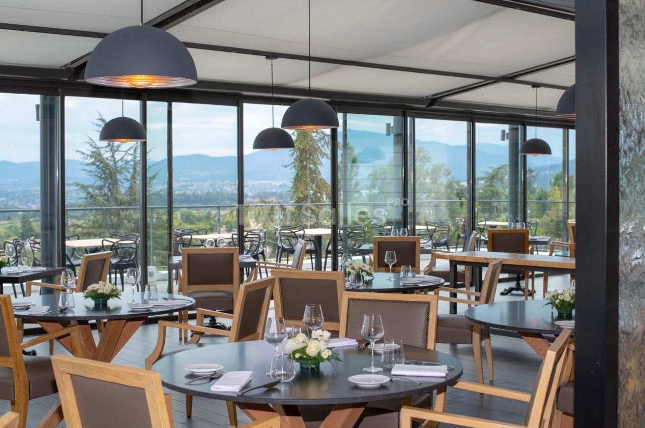 Salle de restaurant lumineuse avec grandes baies vitrées, vue sur un paysage vallonné et tables élégamment dressées.