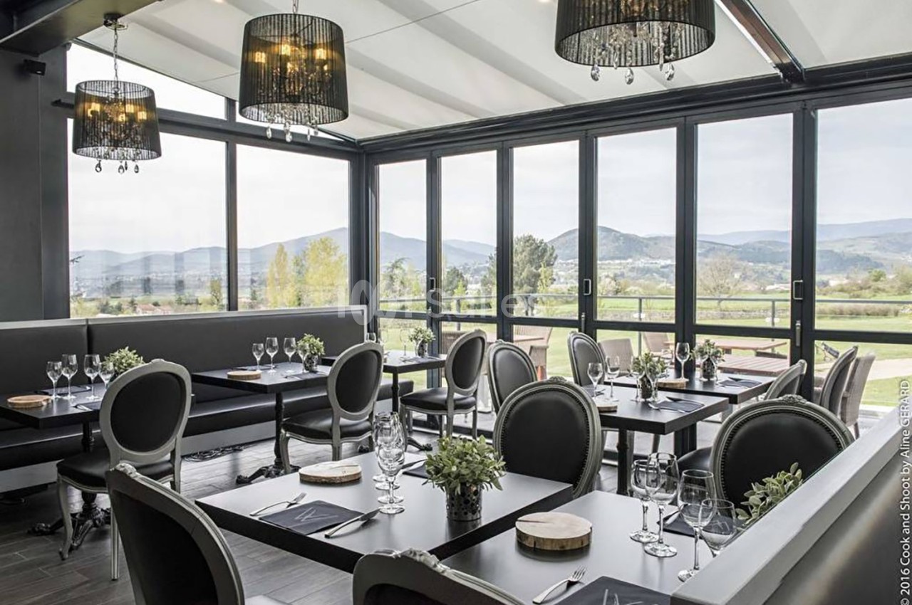 Salle de restaurant lumineuse avec baies vitrées donnant sur un paysage de collines, tables dressées avec élégance.