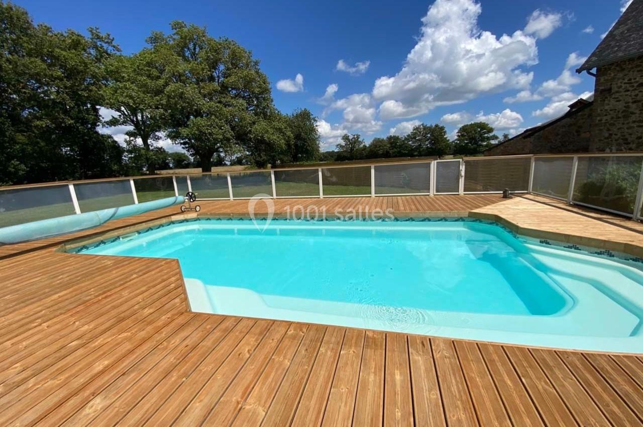 Piscine extérieure entourée d'une terrasse en bois, avec vue sur un jardin arboré sous un ciel partiellement nuageux.