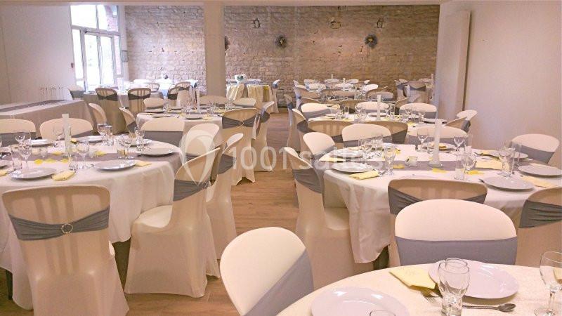 Salle de réception décorée avec des tables rondes dressées, nappes blanches et chaises ornées de rubans gris.