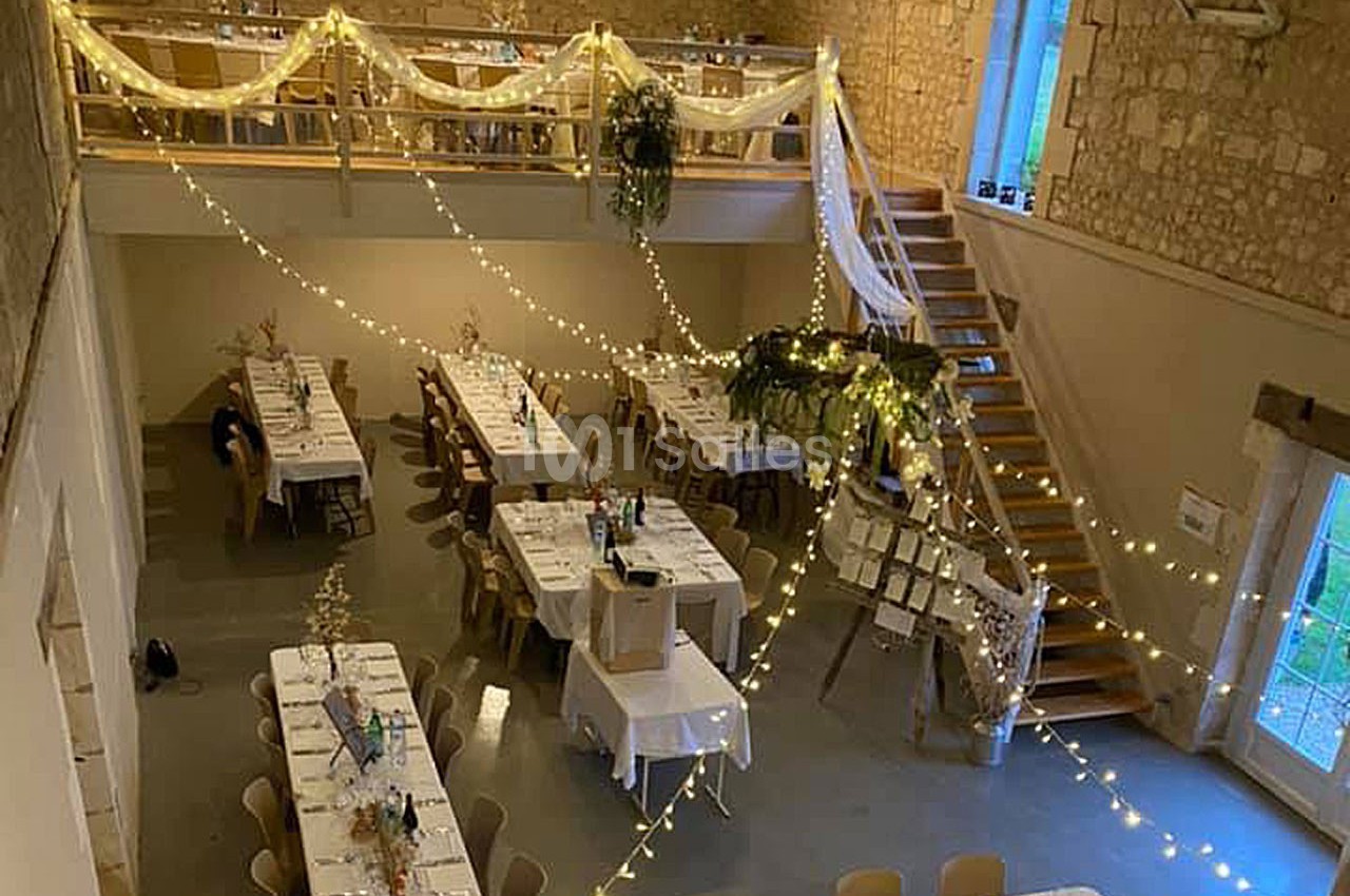 Salle de réception décorée avec des guirlandes lumineuses, des tables dressées et un escalier en bois.