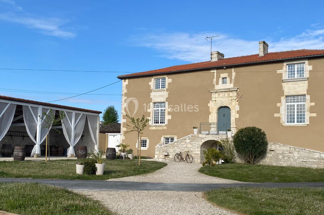 Grande maison en pierre beige avec toit rouge, jardin aménagé, allée gravillonnée et terrasse couverte à gauche.