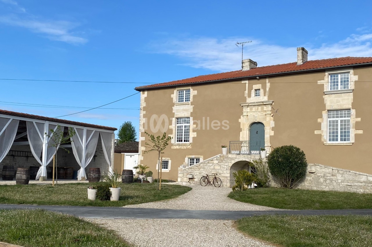 Grande maison en pierre beige avec toit rouge, jardin aménagé, allée gravillonnée et terrasse couverte à gauche.