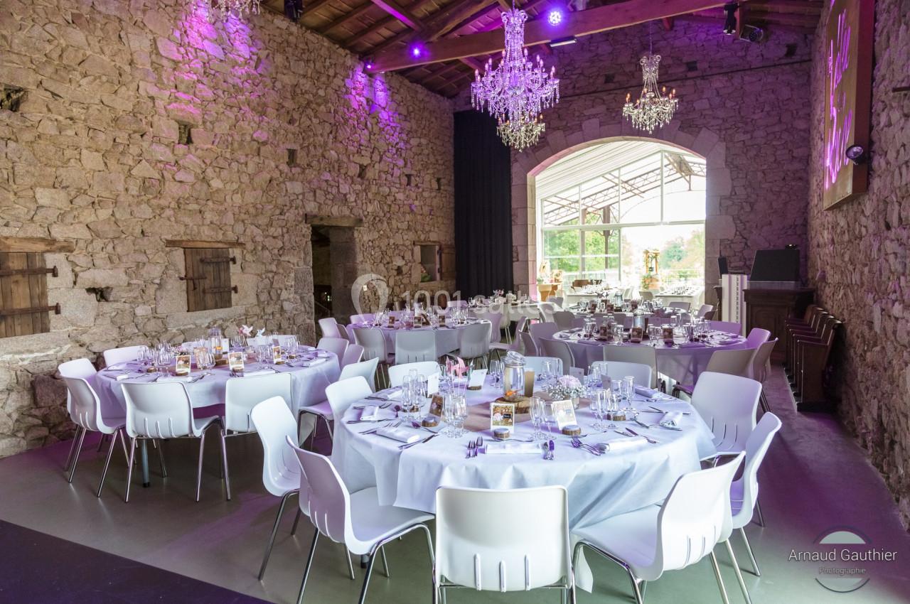 Salle en pierre avec tables rondes dressées pour un événement, éclairée par des lustres et une lumière naturelle.